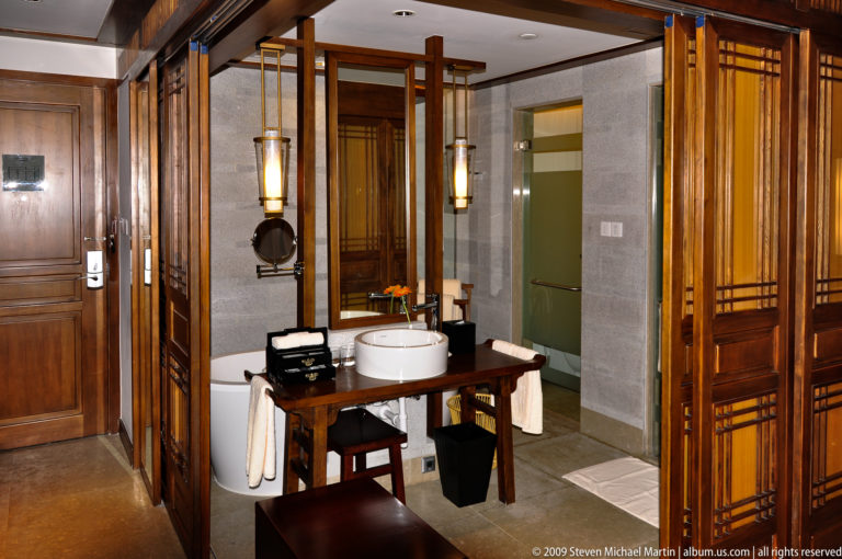 Contemporary hotel bathroom in Lijiang, China, with double wooden vanities, two round white vessel sinks, tall mirrors, and contrasting warm wood and grey stone surfaces.