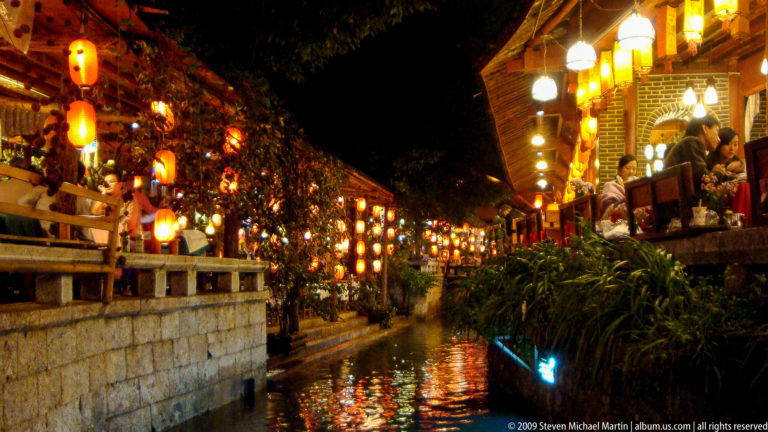 Nighttime view of an illuminated canal in Lijiang, China, with lantern reflections on the water and timber restaurant terraces lining stone canal walls.