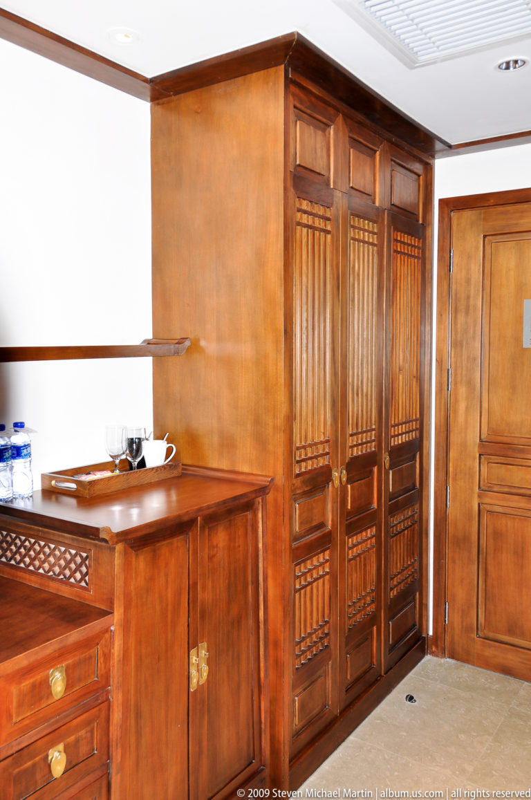 Built-in wooden wardrobe with lattice-front doors above a polished minibar cabinet holding a tray of drinks in a wood-paneled guest room in Lijiang, China.