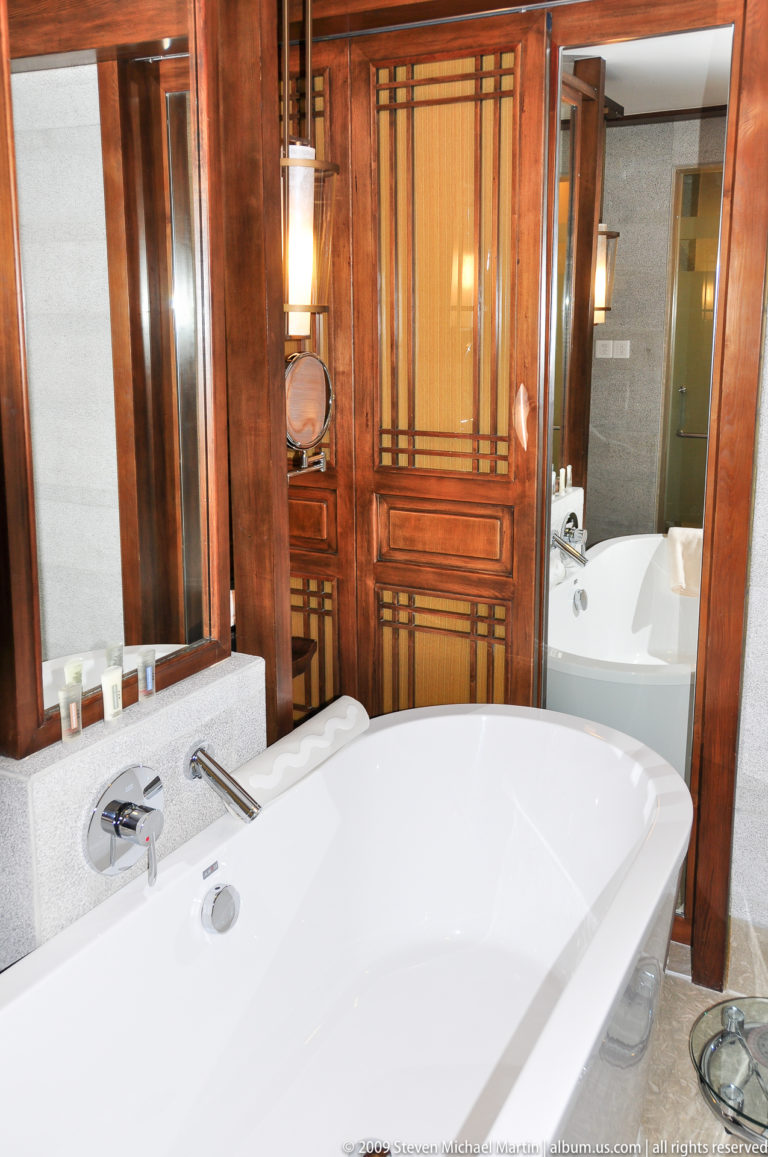 Luxury bathroom in Lijiang, China, with two curved white soaking tubs beside each other and traditional wooden lattice doors set into grey stone walls.
