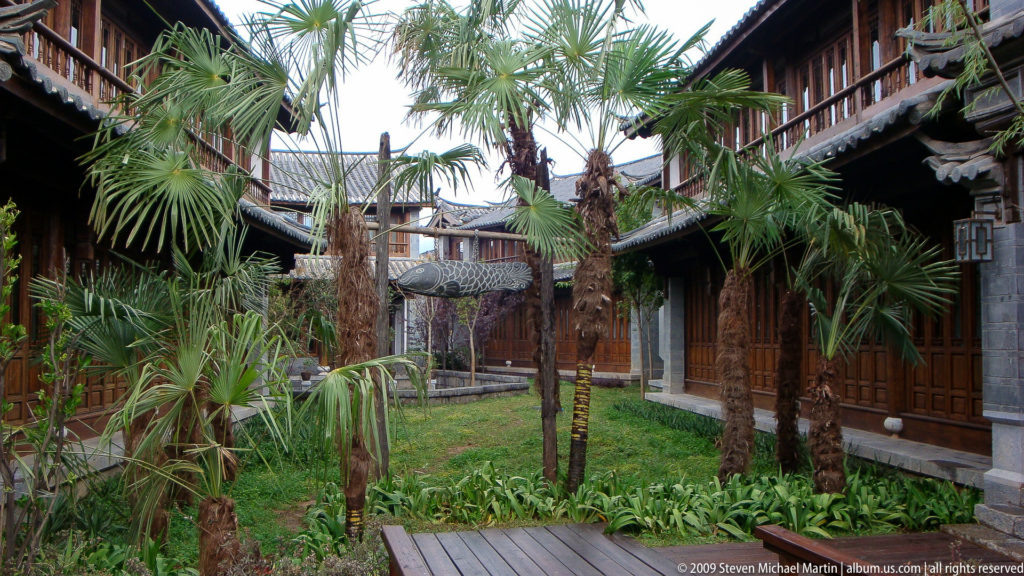 Enclosed Lijiang lodging courtyard with palm trees on a central lawn, surrounded by timber balconies and grey tiled roofs, with a boardwalk and covered garden structure across the center.
