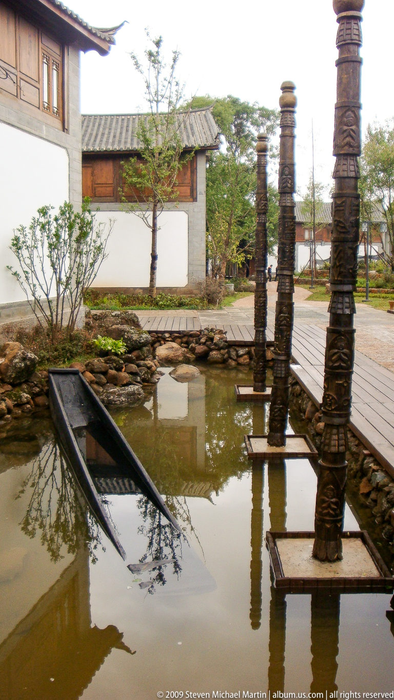 Shallow landscaped pool in Lijiang, China, with tall carved posts bearing stacked mask-like motifs, bordered by boardwalks and rocks.