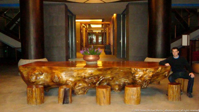 Massive polished natural-root table with an irregular burled wood slab on stump-like supports in a long hotel corridor with dark columns in Lijiang, Yunnan, China.
