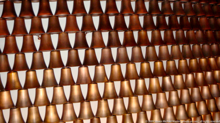 Close-up of a backlit decorative wall in Lijiang, China, showing rows of identical bronze cup-shaped modules with warm light shining through triangular gaps.