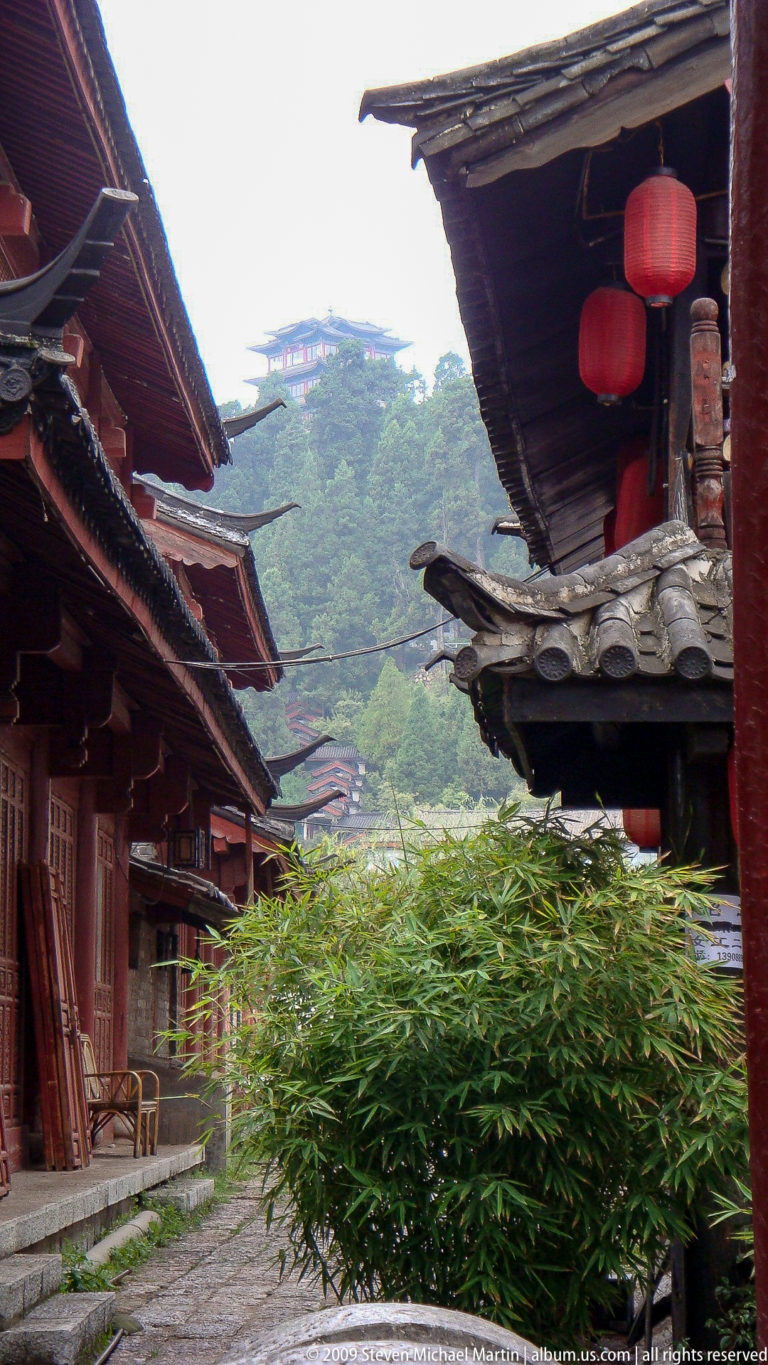 Traditional Lijiang timber buildings with overlapping upturned grey tile eaves framing a bamboo-covered green mountain in the distance.