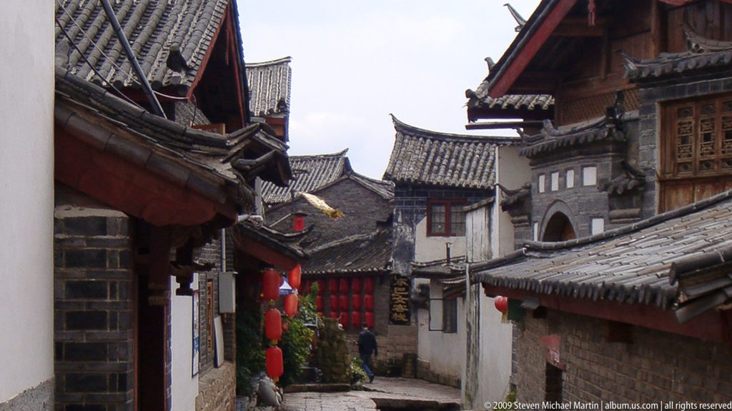 Narrow old town lane in Lijiang, China, with white plaster and grey brick walls, layered tiled roofs, projecting eaves, lanterns, and a small arched doorway.