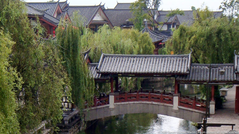 Traditional roofed bridge with grey-tiled canopy and red railings crossing a landscaped pond, with willows and traditional roofs around it in Lijiang, Yunnan, China.