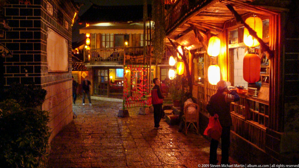 Night view of a stone-paved pedestrian lane in Lijiang, China, lined with lantern-lit timber shops and pedestrians under warm lighting.
