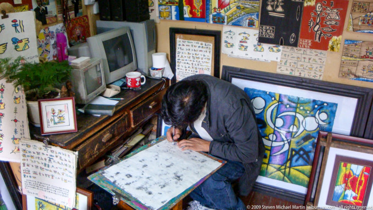 Seated artisan in a small Lijiang studio working with a hand tool on a pale rectangular surface, with pictographic sheets and colorful framed artworks on the walls.
