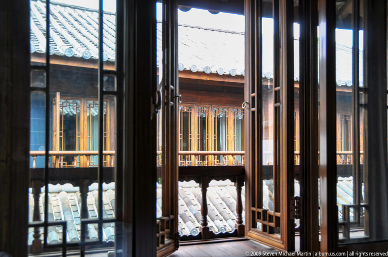 Interior view through open wooden lattice windows toward a sunlit courtyard gallery with a timber façade and grey roof tiles in Lijiang, China.