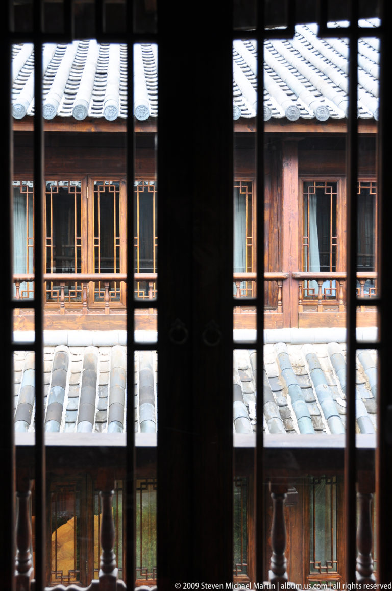 View through dark timber doors with circular metal pulls toward an opposite gallery with wooden balcony rail and grey tiled roof in a traditional Lijiang courtyard.