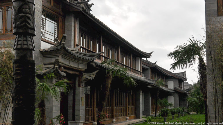 Traditional-inspired resort guesthouse wing in Lijiang, Yunnan, China with timber balcony, tiled eaves, central carved entry canopy, and wood-framed windows beside landscaped palms.