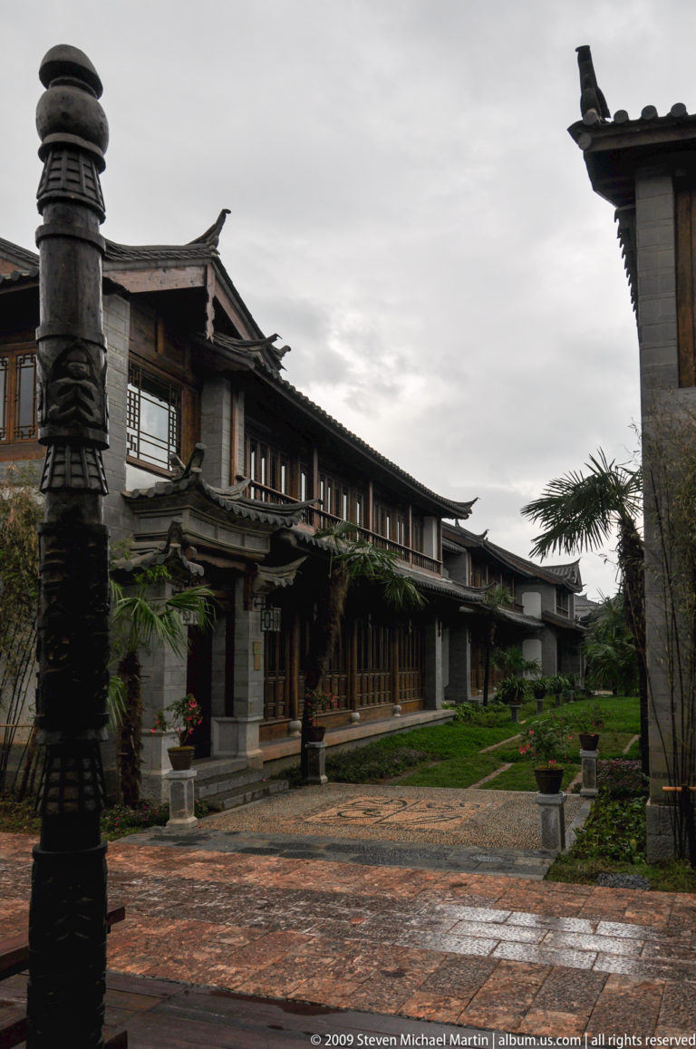 Two-story timber-and-stone guesthouse in Lijiang, Yunnan, with traditional roof forms, an upper-level wooden balcony with lattice windows, and palms and garden beds along the façade.