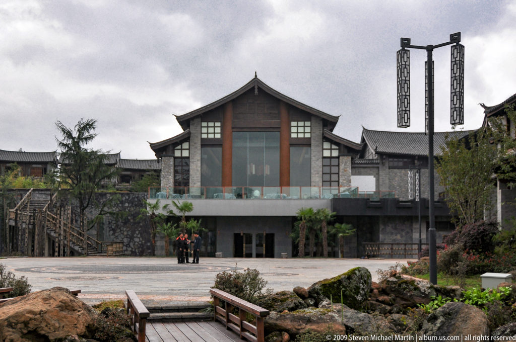 Large contemporary stone-and-timber building in Lijiang, China, with a broad glazed central façade under a gabled roof and a landscaped plaza with decorative lamp posts in front.