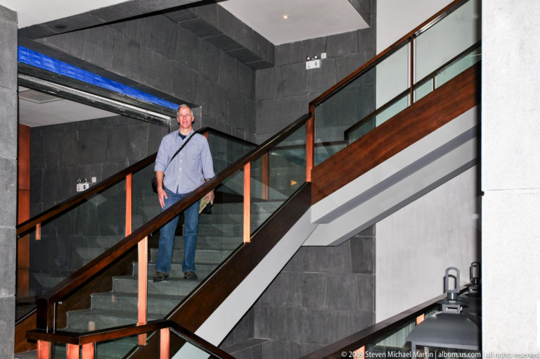 Male visitor standing midway on stone steps of a modern hotel or resort stairway with glass balustrade and dark stone walls in Lijiang, Yunnan, China.