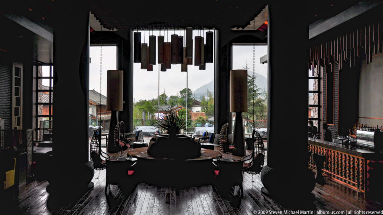 Dark timber restaurant dining room in Lijiang, China, with a formally set round table, high-backed chairs, suspended rectangular lights, and mountains visible outside.