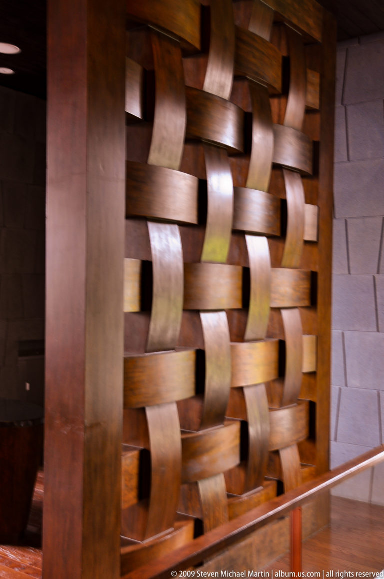 Close-up of broad, curved wooden strips in a polished basket-weave screen against a pale masonry wall in an interior in Lijiang, Yunnan, China.