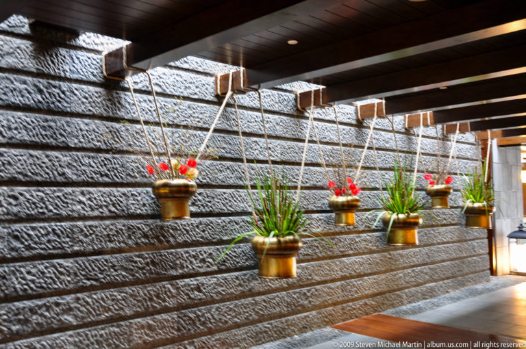 Row of small brass pots with grass-like plants and red decorations hanging on cords against a textured stone wall in an interior corridor.