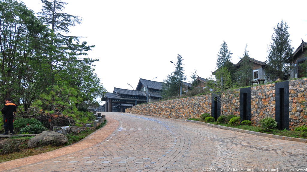 Curving paved drive with a stone retaining wall leading uphill to dark-timbered traditional-style buildings and landscaped trees in Lijiang, Yunnan, China.