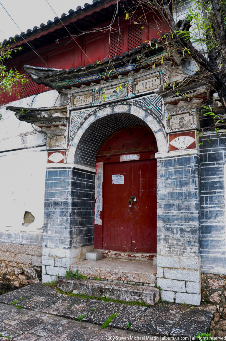 Decorated blue-grey brick arch with painted geometric and floral ornament and tiled edging surrounding closed red double doors in Lijiang, Yunnan, China.
