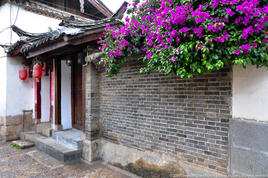 Traditional Lijiang courtyard wall and doorway in China, with dense magenta bougainvillea spilling over the roofline above grey brick, white plaster, red lanterns, and stone paving.