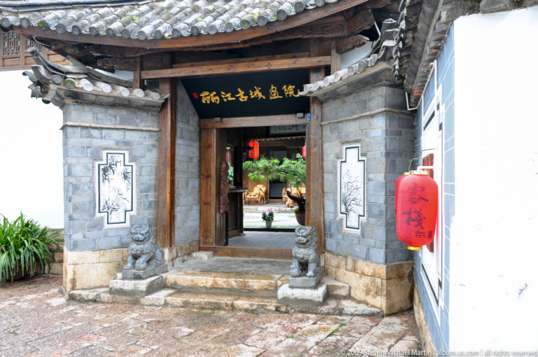 Grey-brick and timber courtyard doorway in Lijiang, China, with a black signboard bearing gold Chinese characters, a red lantern, and two small stone guardians at the entrance.