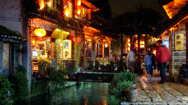Small water channel beside a stone walkway and lantern-lit traditional timber shopfronts with potted flowers in Lijiang, Yunnan, China.