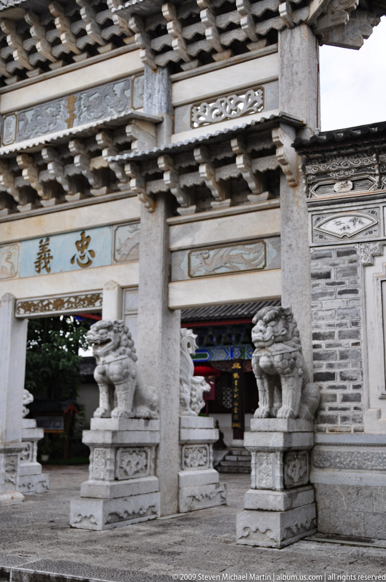 Close view of a traditional Lijiang stone entrance in Yunnan, China, with gold-toned Chinese characters on a pale panel above two stone lion guardians under carved lintels.