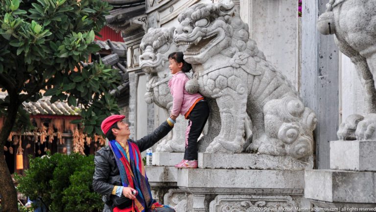 Man lifting a child onto a guardian-lion pedestal as the child reaches toward the carved stone figure, with large stone lions and a gateway wall in the background in Lijiang, China.