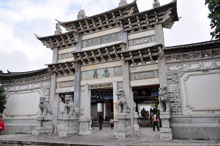 Ornate stone paifang gateway in Lijiang, Yunnan, with layered carved brackets, guardian lions, and Chinese character panels around the central entrance.