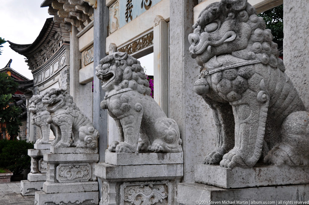 Three carved stone guardian lions on decorated pedestals in front of an ornate ceremonial gateway in Lijiang, Yunnan, China.
