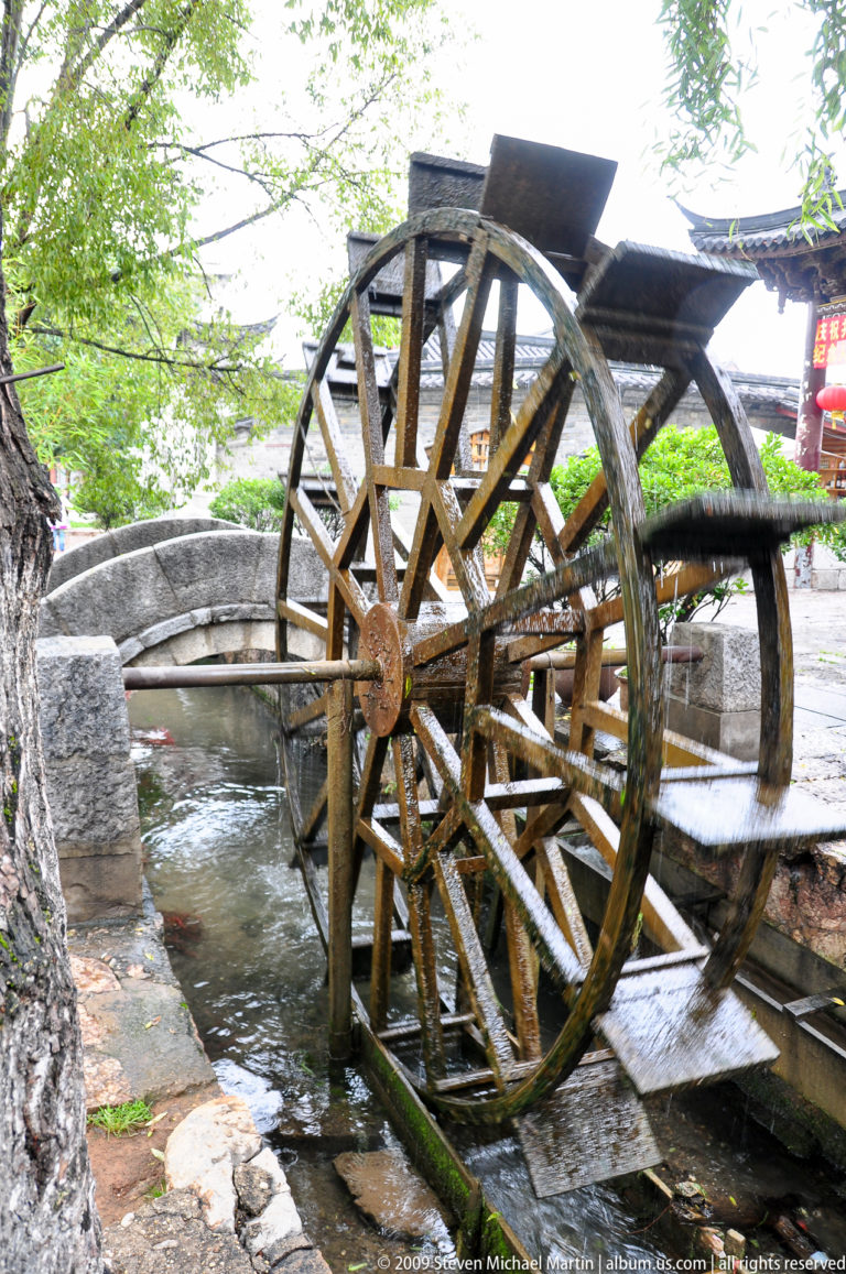 Side view of a wooden waterwheel turning in a stone channel, with wet paddles blurred in motion between a stone bridge and water basin in Lijiang, Yunnan, China.