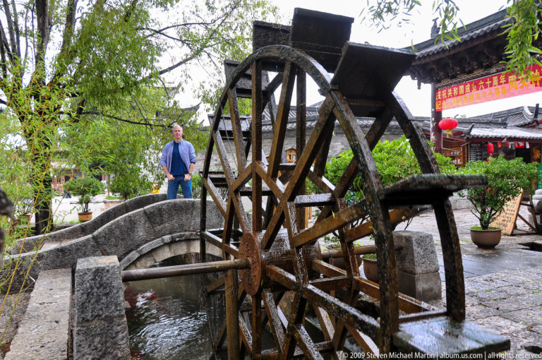 Traditional wooden waterwheel in a channel beside a small stone bridge with a male visitor standing on the bridge in Lijiang, Yunnan, China.