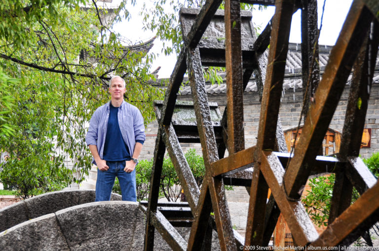 Male visitor standing behind the spokes of a wooden wheel, with a stone bridge and timber building in the background in Lijiang, China.