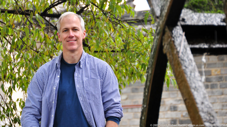 Male visitor in a blue shirt and checked overshirt standing beside a waterwheel framed by willow branches in Lijiang, Yunnan, China.