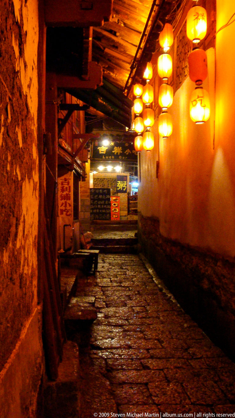 Wet stone-paved lane in Lijiang Old Town at night, rising between plastered walls and lit by warm shop lanterns with Chinese signs.