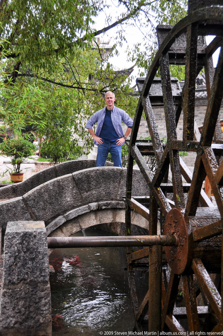 Male visitor with hands on hips standing behind a small arched stone bridge and large dark timber wheel in Lijiang, Yunnan, China.