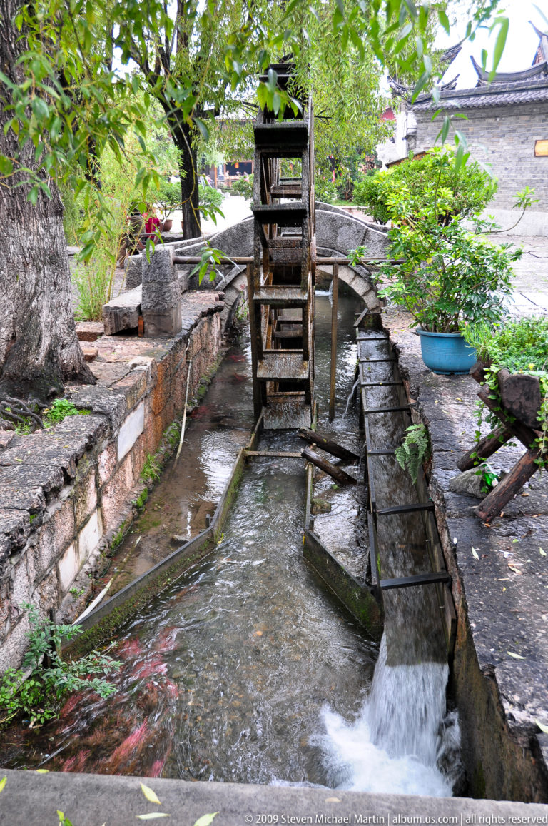Small wooden waterwheel in a narrow stone-lined water channel in Lijiang, Yunnan, China, with paddles dipping into flowing water and masonry walls and potted plants along the sides.