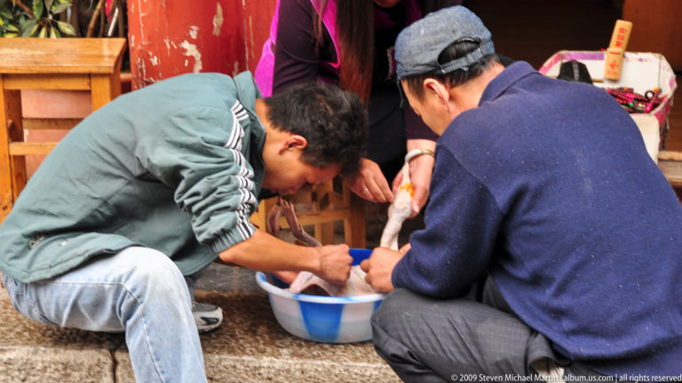 Two men crouch over a blue basin preparing food at a street stall in Lijiang, Yunnan, China, with a shop counter and another person standing behind them.