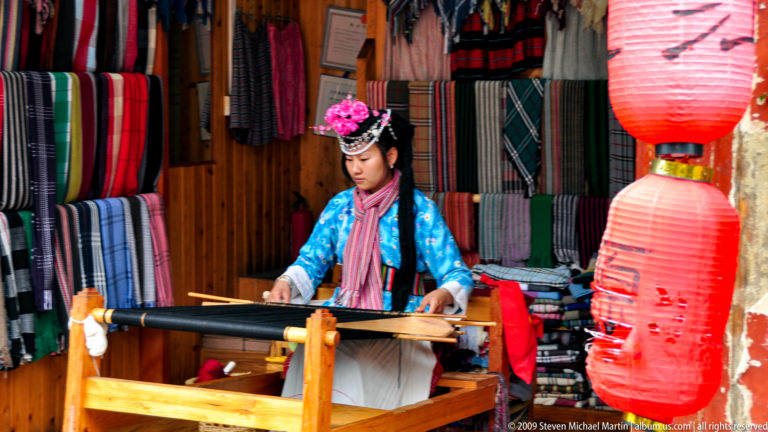 Young woman in a bright blue traditional-style blouse and decorative headpiece weaving on a hand loom in a textile shop in Lijiang, Yunnan, China.