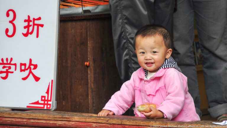 Toddler in pink standing behind a wooden rail, smiling and holding food, with adult legs and a sign with red characters behind.