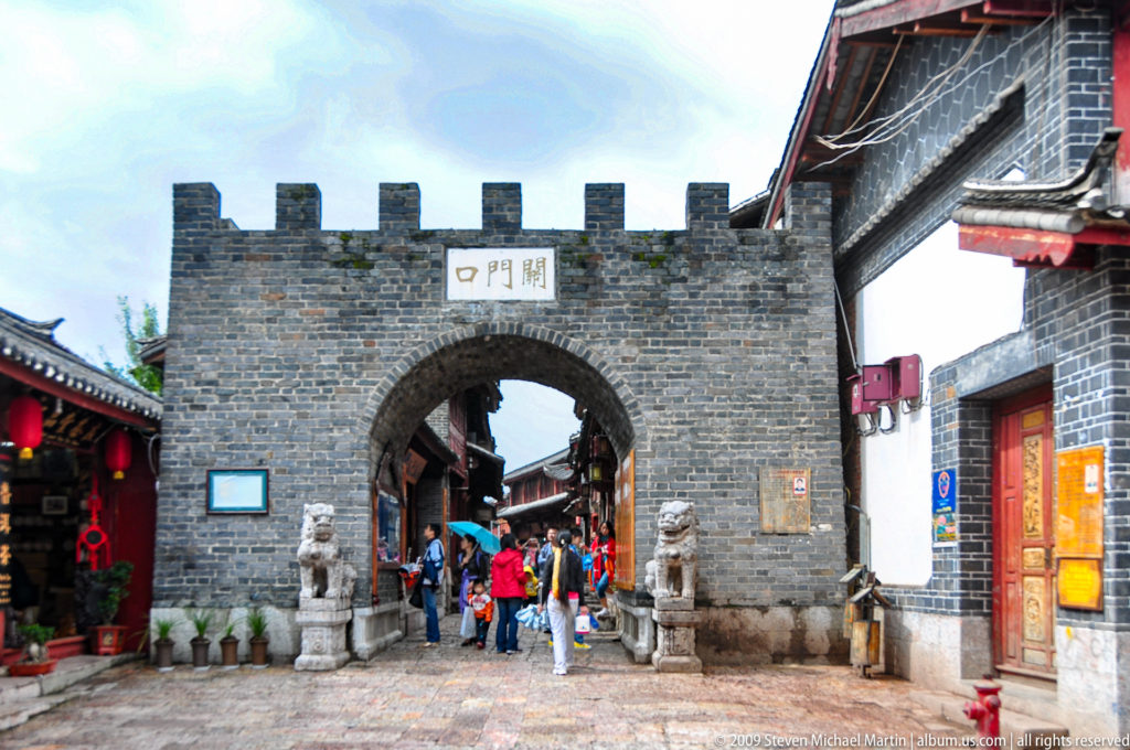 Grey-brick crenellated gateway with a round arched entrance, stone lion guardians, and people walking into a pedestrian district in Lijiang, Yunnan, China.