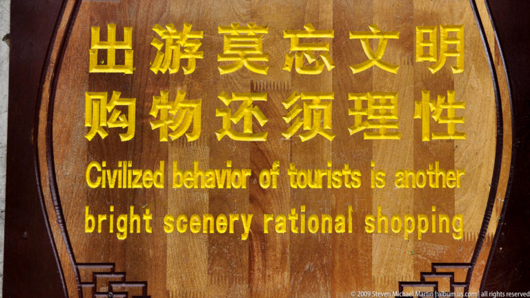 Bilingual public sign in Lijiang, Yunnan, with gold Chinese characters above English text on a dark wooden panel encouraging civilized behavior and rational shopping.