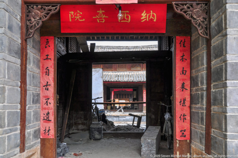 Traditional Lijiang courtyard gateway with timber lintels, grey brick walls, and red vertical calligraphy couplets opening onto successive courtyards.