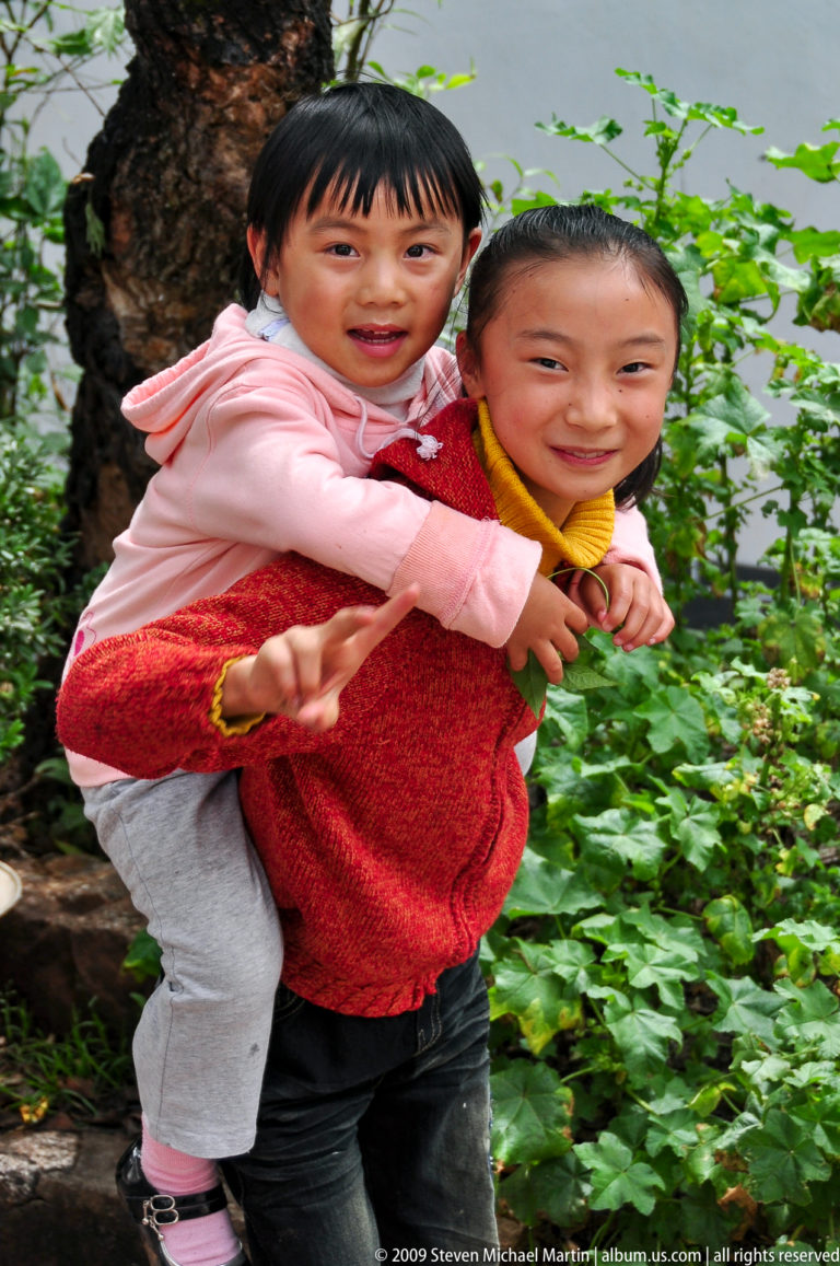 Two girls outdoors in Lijiang, China, one giving the other a piggyback ride and making a peace sign in front of green foliage.