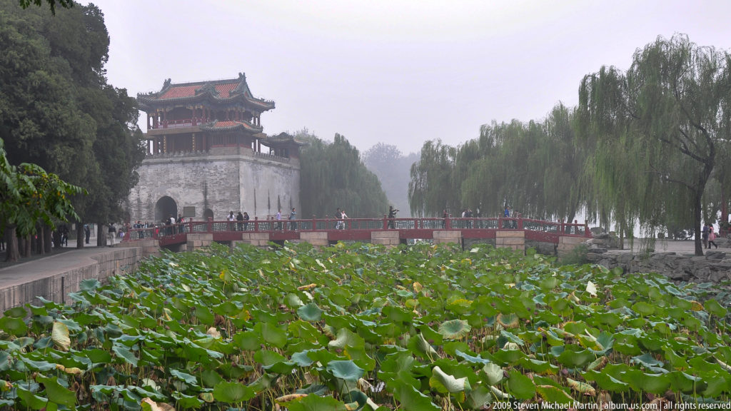 Broad lotus pond with a red bridge and gray pavilion-topped tower beyond, framed by willow trees at the Summer Palace.