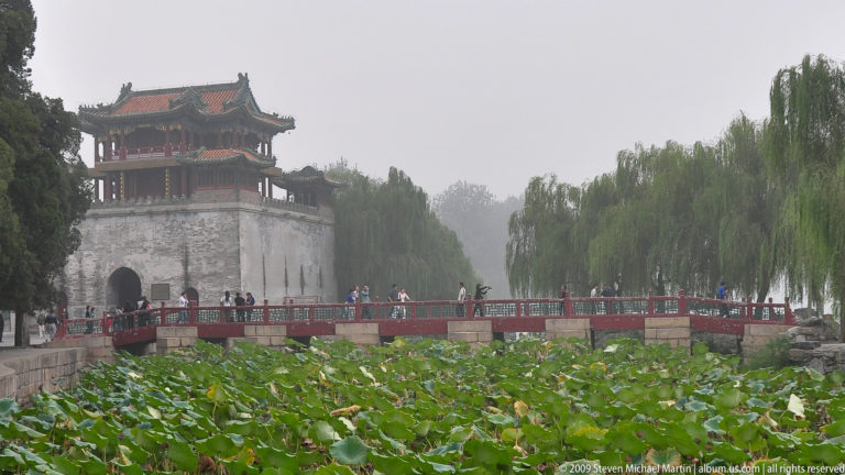 Gray pavilion-topped tower beside a red-railed bridge and dense lotus pond, with willow trees in the hazy Summer Palace garden.