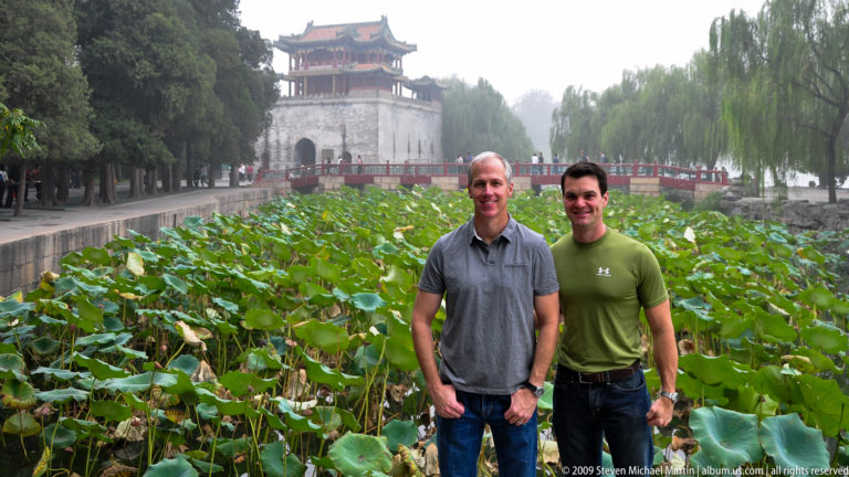 Two men beside a dense lotus pond with a red bridge, willow trees, and a gray pavilion-topped tower in the background.