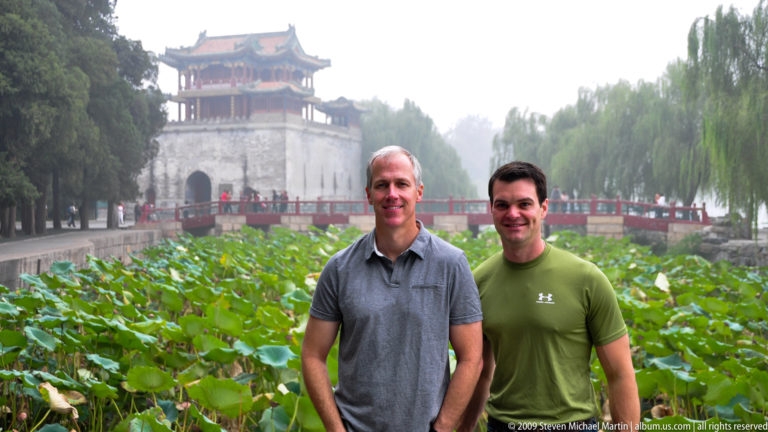 Two men standing before a lotus pond, with a gray tower, red-railed bridge, and willow trees behind them at the Summer Palace.