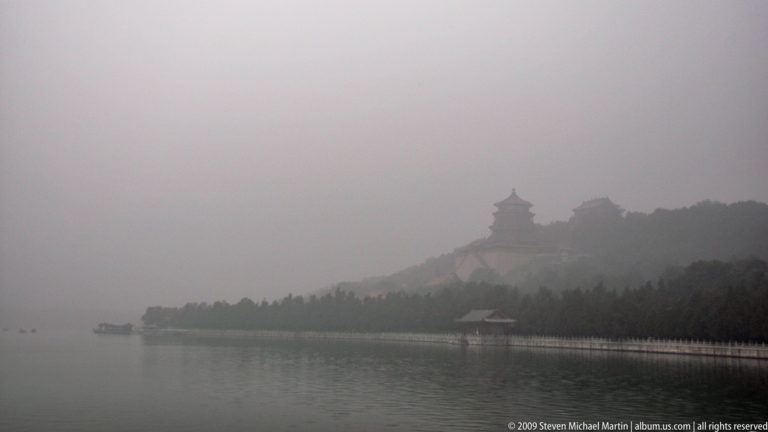 Misty Kunming Lake with the wooded silhouette of Longevity Hill, faint palace buildings, and a small pavilion along the distant shore.